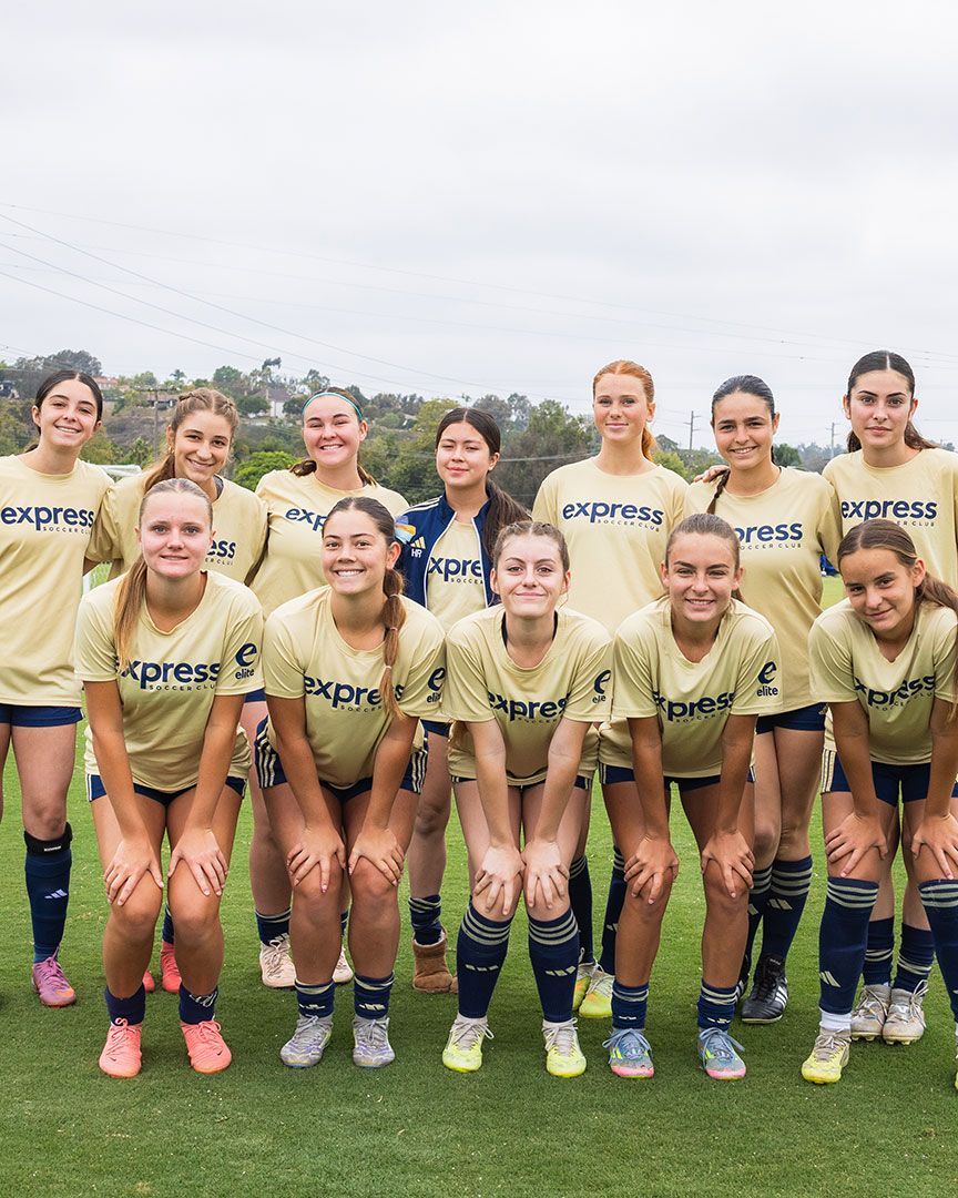 Soccer team posing on a grassy field, wearing gold jerseys and navy socks, smiling for a group photo