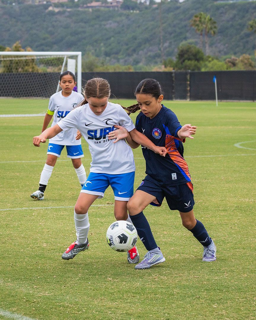 Two youth soccer players battle for the ball on a grass field, with a goal and hills in the background.