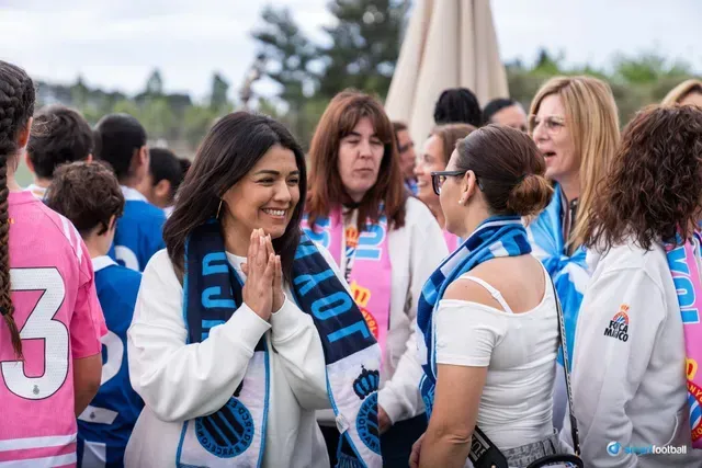 Woman with scarf greets a group of people outdoors. Supporters are wearing blue and pink jerseys.