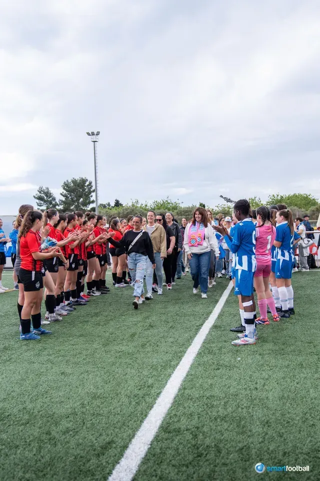 Soccer teams in red and blue lines greet others on the field, spectators walk between them.