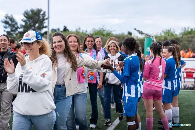 People clapping as female soccer players in blue and pink uniforms walk onto a field.
