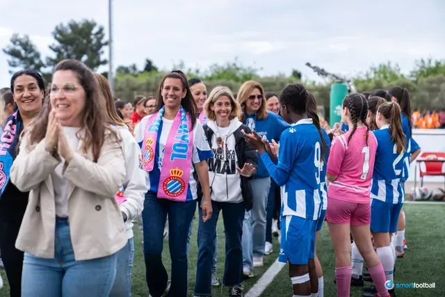 Women clapping, smiling, and lining up with soccer players on a green field.