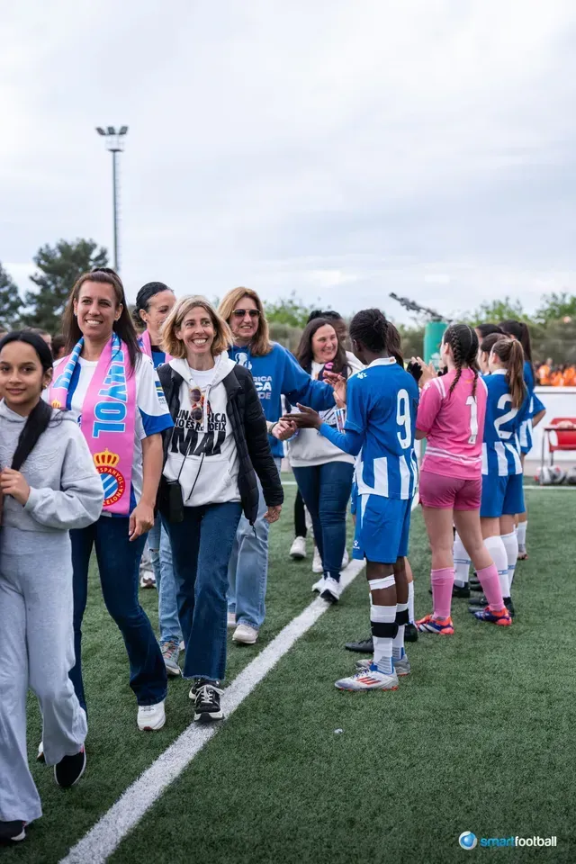 Soccer players in blue & pink uniforms, shaking hands with smiling women on a green field.