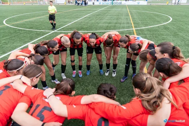 Soccer team in red jerseys huddles on a green field, referee watches.