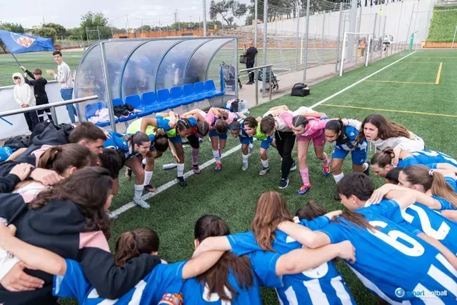 Soccer team in blue and pink jerseys huddles on the field, arms around each other.