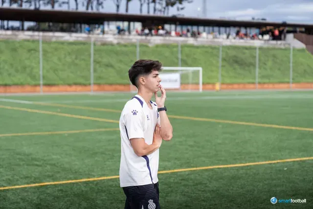 Young man on a soccer field, looking thoughtful, wearing a white polo shirt with blue accents.