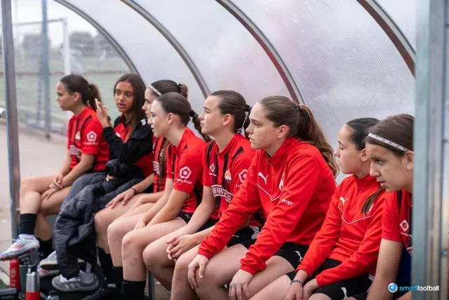 Girls' soccer team in red jerseys sit on a bench, watching a game.