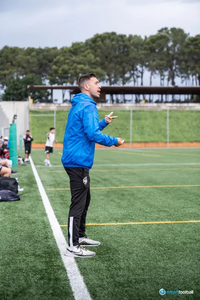 A man in a blue jacket gestures on a soccer field, coaching players during a game.