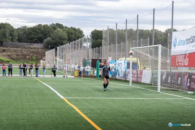 Soccer player kicks ball into a net on a green field; spectators watch.