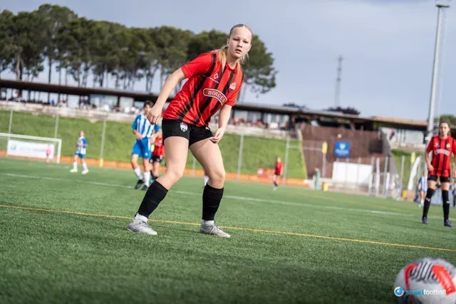 Female soccer player in red and black uniform on a green field, looking towards the ball.