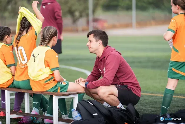 A soccer coach kneels to talk with young players wearing orange/green uniforms. They sit on a bench, field in background.