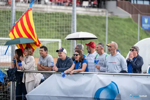 Spectators watch a sporting event, some holding umbrellas. A Catalan flag waves.