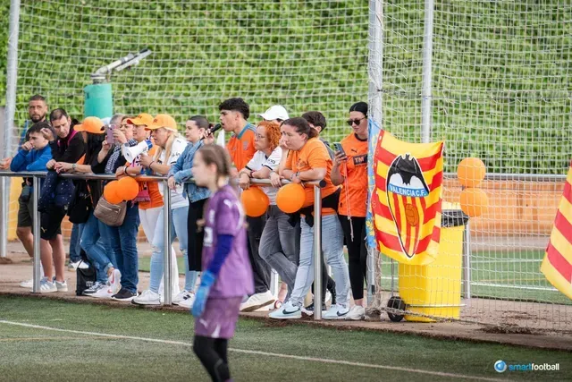 Spectators in orange cheer at a soccer game, some holding balloons and a flag.
