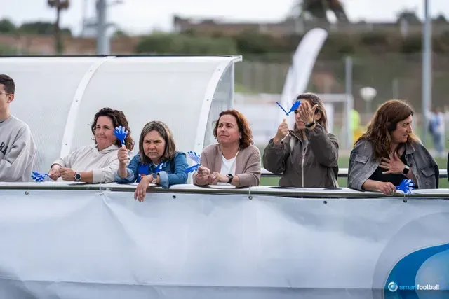 Spectators at a soccer game cheering, holding blue noisemakers. They are outside, under a shelter.
