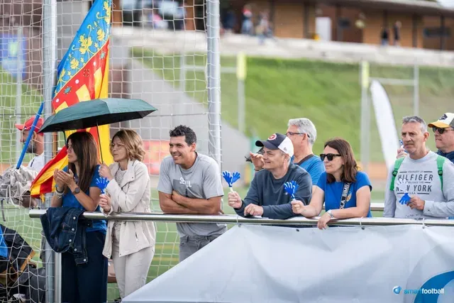 Spectators at an outdoor event, some holding blue items and a Valencia flag, watching.
