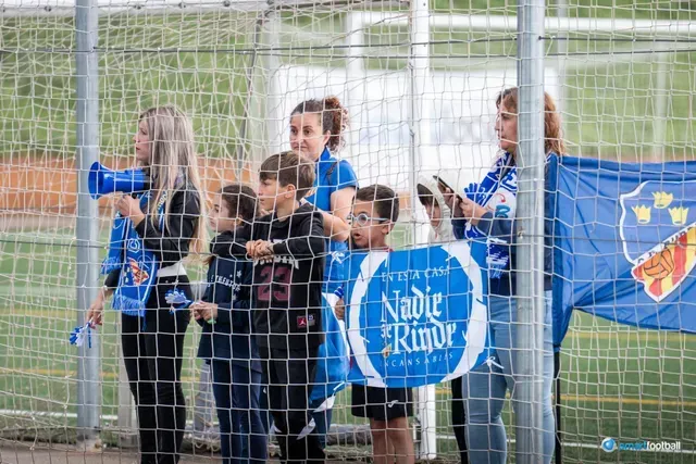 Fans cheering at a soccer game behind a fence; one woman with megaphone, others holding a banner.