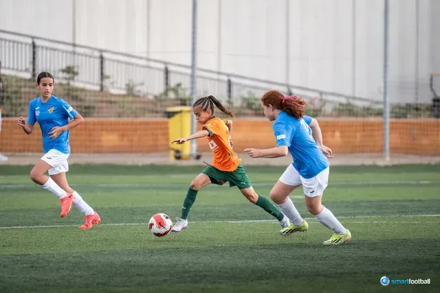 Soccer players on a green field, one in orange dribbles the ball, two in blue chase.