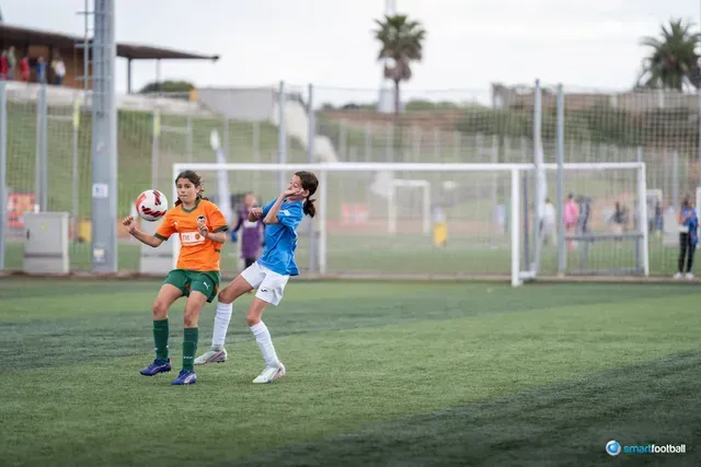 Two female soccer players in action on a green field. One in orange heads a ball; the other in blue jumps to compete.