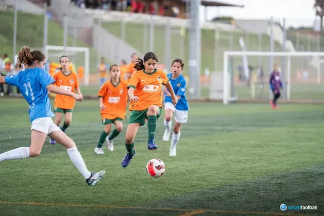Soccer players, mainly girls, in action on a green field. Orange and blue team uniforms. One girl kicks the ball.