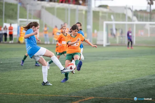 Two girls in a soccer match: one in blue, kicking at the ball; the other in orange, dribbling it. Green field.