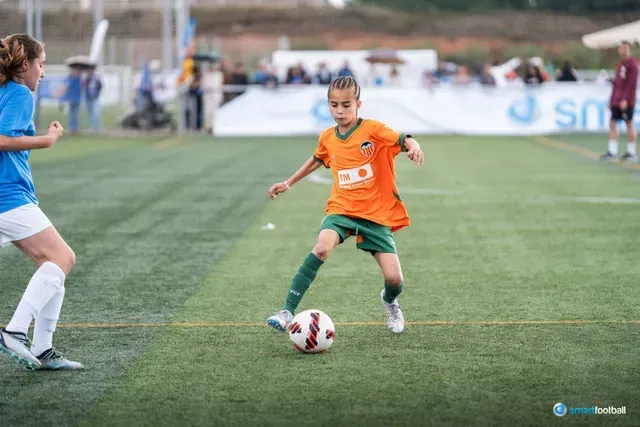 Boy in orange jersey dribbling soccer ball on green field; girl in blue defending.