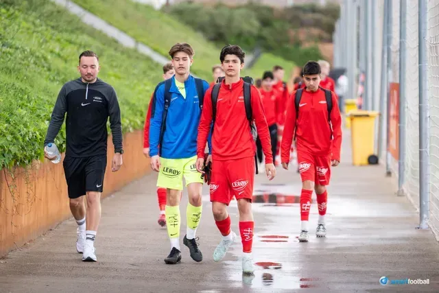 Soccer team in red and blue uniforms walks on a path, led by a coach.