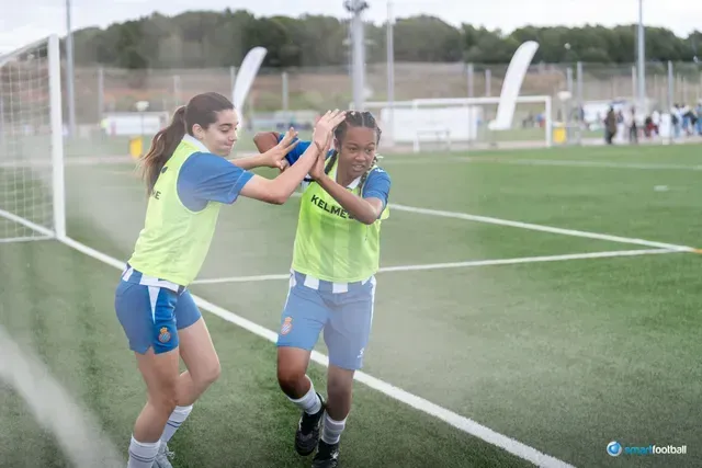 Two women in soccer uniforms high-five on a green field. One wears a blue shirt with a green stripe.