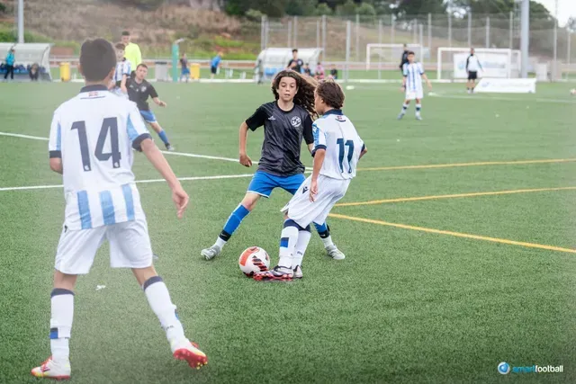 Youth soccer players in action on a green field. Two players face off over the ball; others watch.