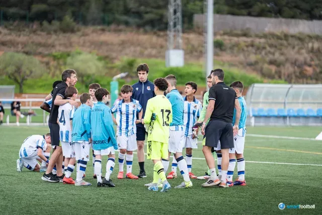 Youth soccer team huddle on a green field with coaches. Players in blue and white jerseys, some are listening.