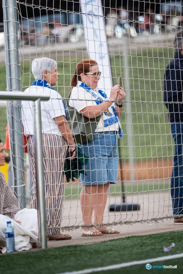 Two women at a soccer game, one taking a photo through the fence.
