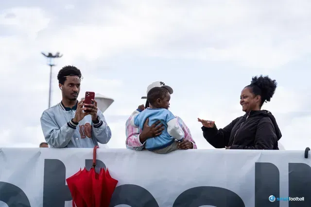 Man taking photo, woman holding child, another woman smiles. Cloudy sky background.