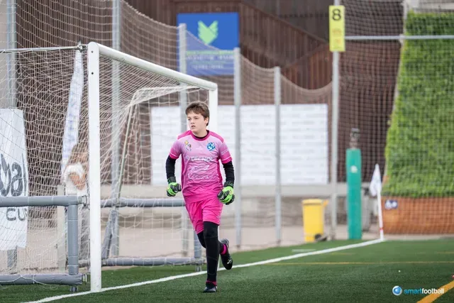 Young soccer goalie in pink uniform running on a green field, near the goal.