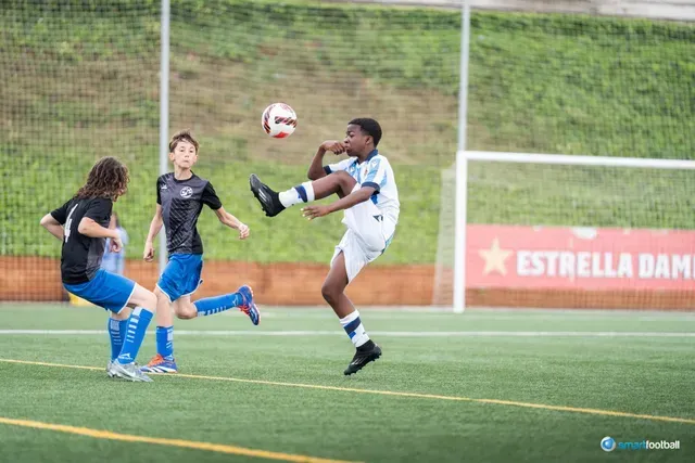 Boy in white kicks soccer ball mid-air during a game. Two other players are nearby on the green field.