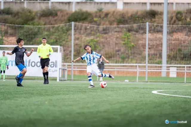 Soccer player kicking the ball on a green field. Another player and referee are nearby.