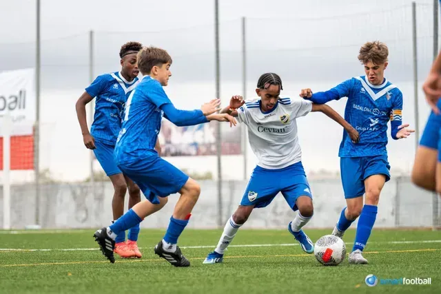 Soccer team in red and white checkered jerseys on a green field. Players are interacting, some looking on.