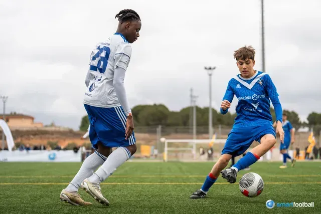 Two young soccer players in blue uniforms on a green field, one dribbling the ball, the other defending.