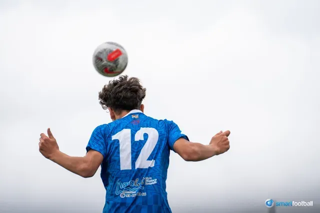 Soccer player in blue jersey, number 12, heading a ball up in the air against a cloudy sky.