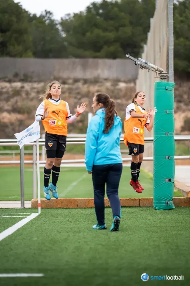 Three young female soccer players in orange practice drills with a coach on a green field.