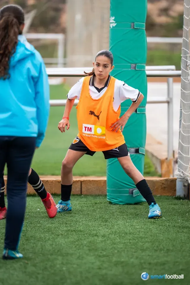 Girl in orange vest on a soccer field, legs apart, hands on hips, with another person in blue nearby.