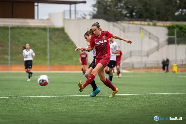 Woman in maroon jersey kicking soccer ball on a green field, another player nearby.