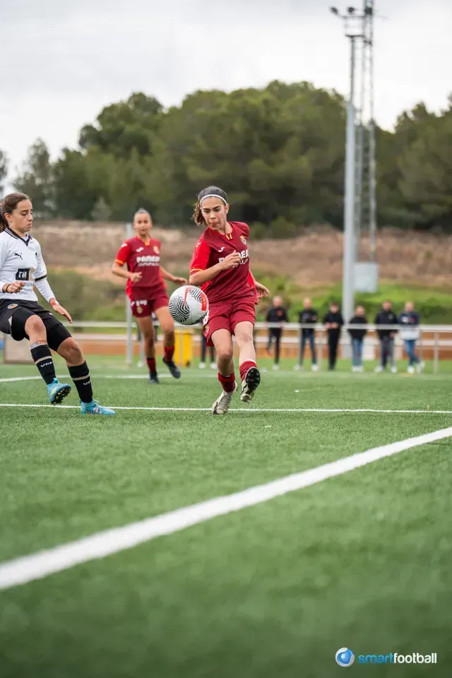 Soccer player in burgundy kicks the ball, another player defends; green field, outdoor game.