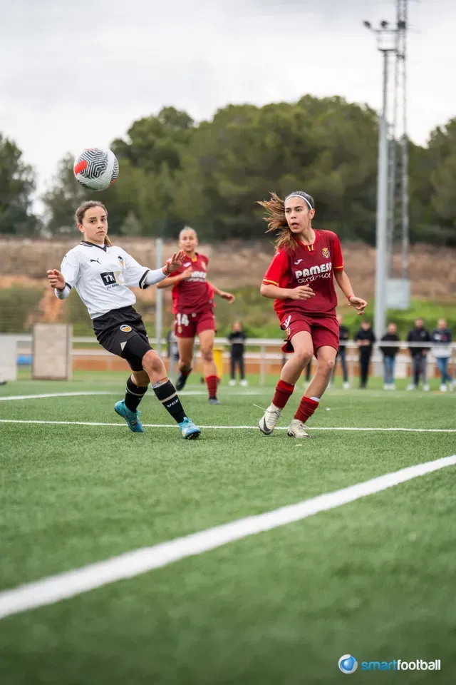 Soccer players on a green field. A player in blue attempts to tackle a player in white, with a referee nearby.