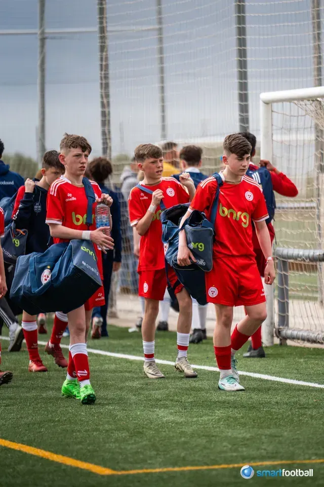 Soccer team in red jerseys walking on a green field, carrying bags. Cloudy day.