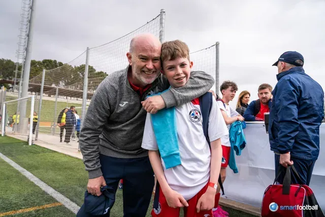 Man with arm around boy on soccer field, smiling. Both in sports attire, with spectators in the background.