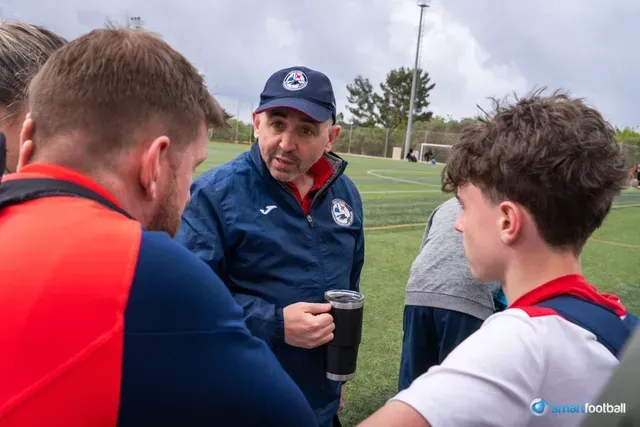 Coach in blue jacket and cap speaks to soccer players on a green field.