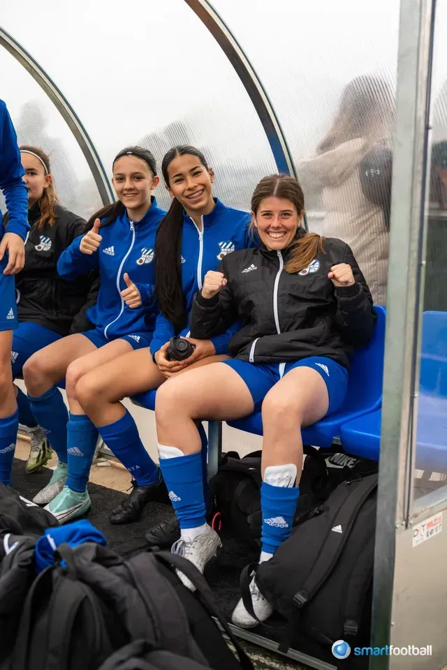 Female soccer players in blue uniforms and jackets on the bench, smiling and making fist pumps.