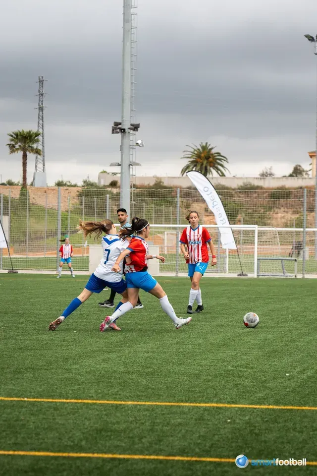 Two women playing soccer on a green field, one kicking the ball, cloudy sky.