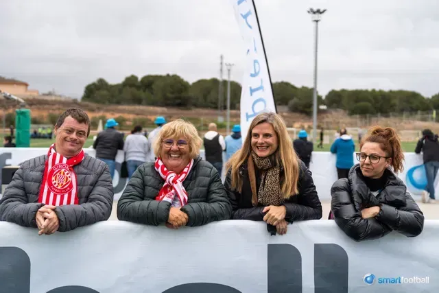 Four people smiling, leaning on a barrier, wearing scarves and jackets, outdoors at a sports event.