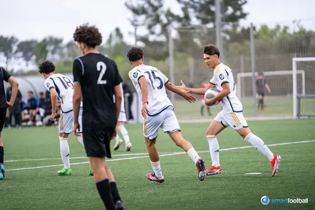 Soccer players on a field; player with ball high fives another, wearing white and gold uniforms.
