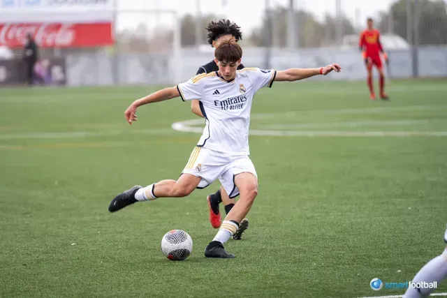 Soccer player in white jersey kicking ball on green field, another player in black close behind.